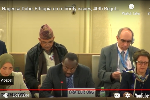 [ai] A panel of speakers at a conference, with a focus on a man speaking into a microphone. In the background, individuals are seated, including a woman wearing a hijab and a man with glasses. The setting appears to be a formal meeting.